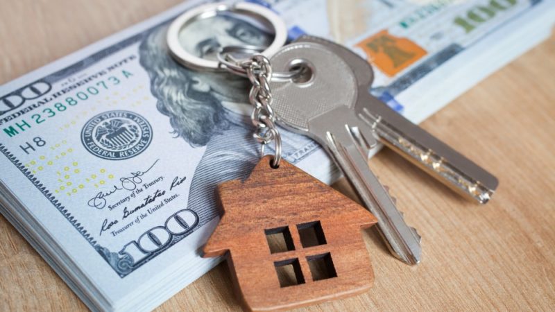 A set of keys with a keychain shaped like a house sits atop a stack of 100 dollar bills.