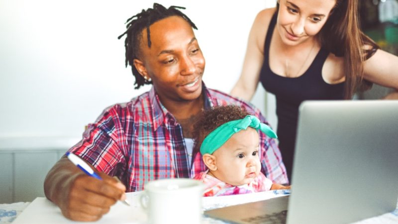 A man writes notes with a baby on his lap and a woman at his shoulder. They are all looking at a laptop.
