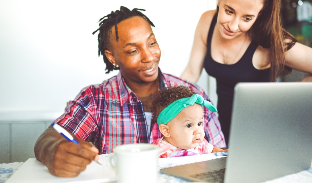 A man writes notes with a baby on his lap and a woman at his shoulder. They are all looking at a laptop.