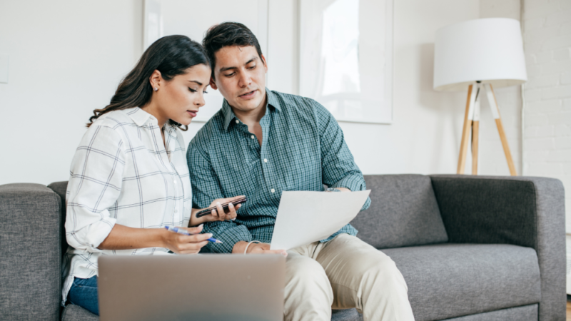 A woman and man sit on a couch looking over a document. A laptop computer is in the foreground