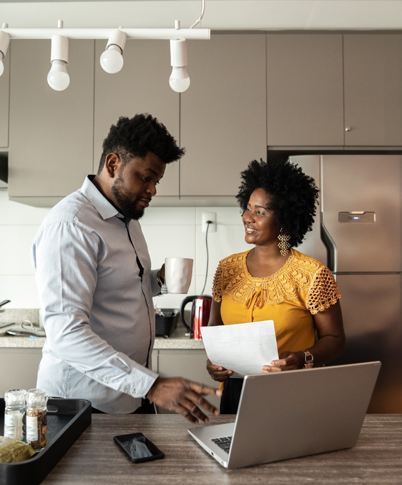 A man and woman stand in their kitchen and share information on their laptop and forms