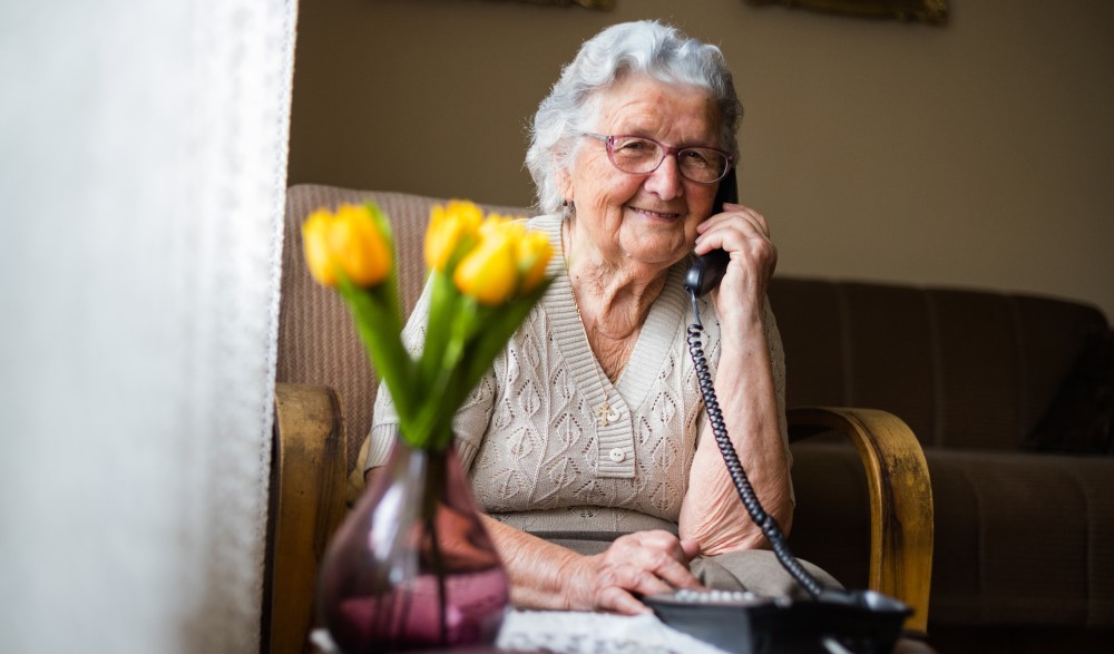 Ano older adult woman holds a classic corded telephone to her ear while seated in a chair with yellow flowers on a nearby table