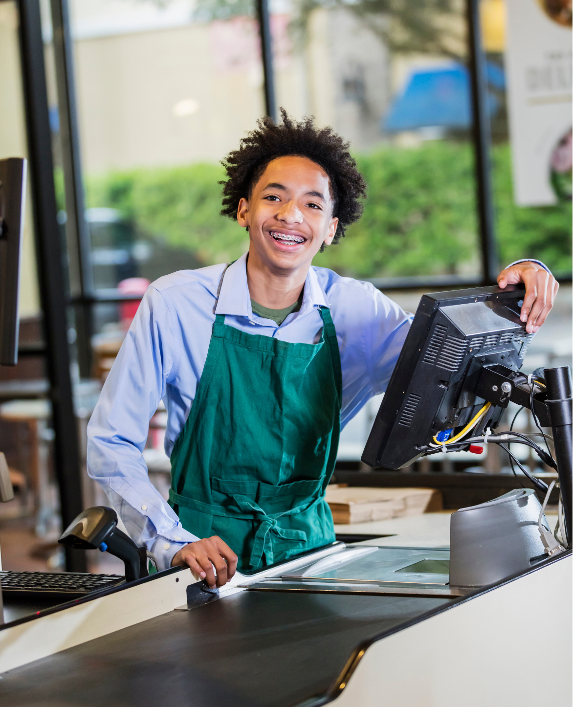 A young man with a green apron smiles at his job as a grocery store cashier