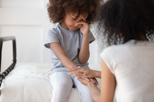 A young child with brown curly hair wipes her eye as a woman with brown curly hair holds her hand in a comforting manner
