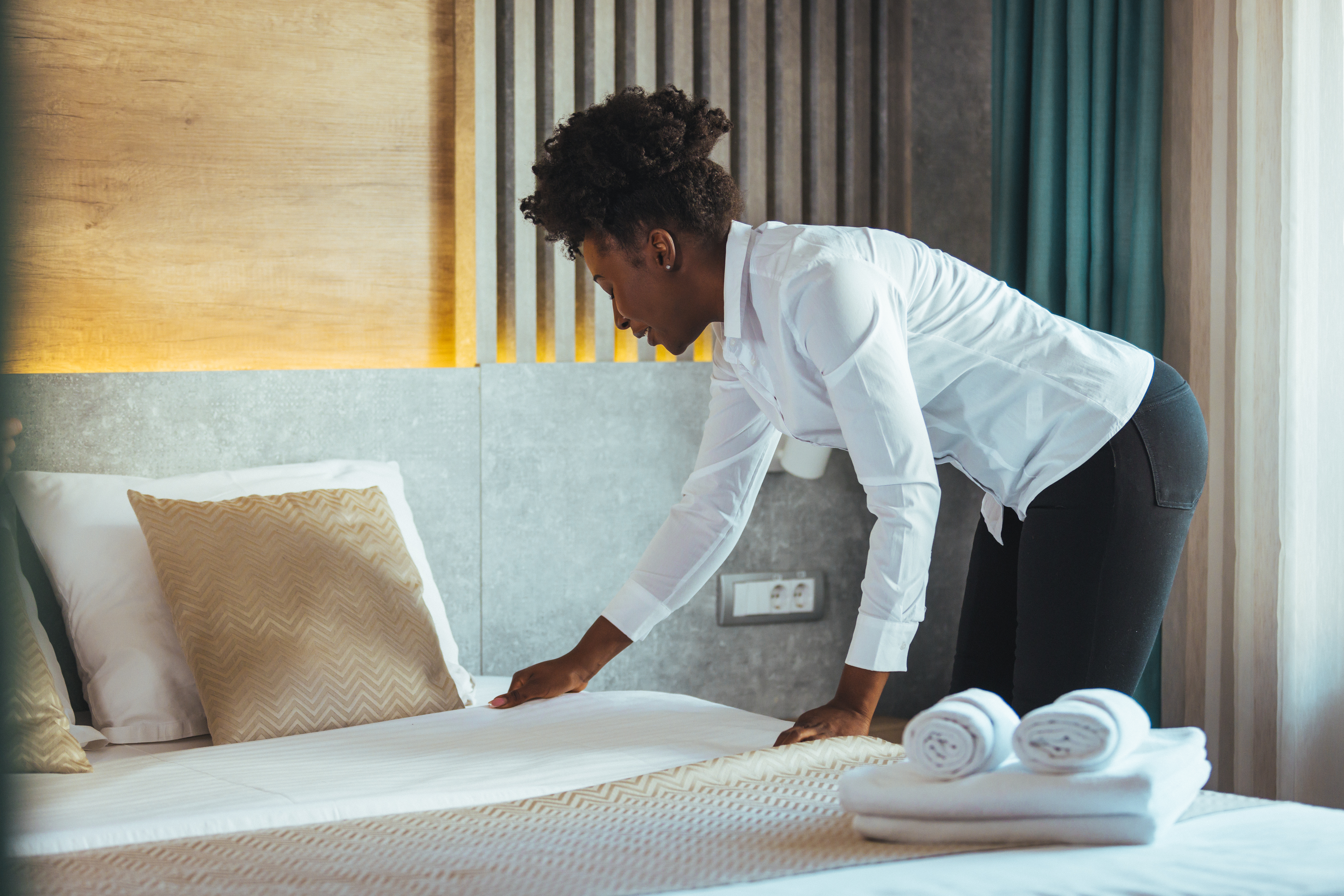 A young African American hotel worker in business casual attire smooths out the covers of a hotel bed.