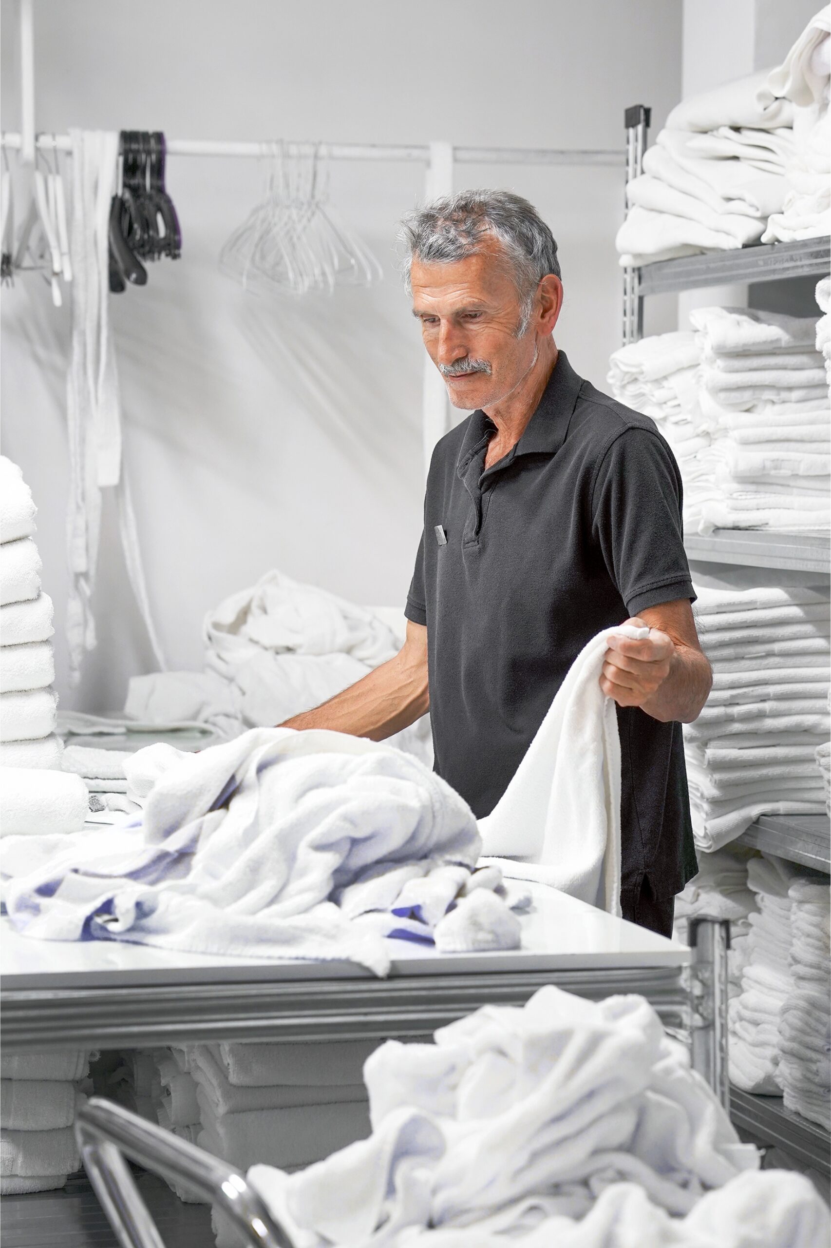 An older Caucasian man with a nametag folds hotel towels in a laundry room.